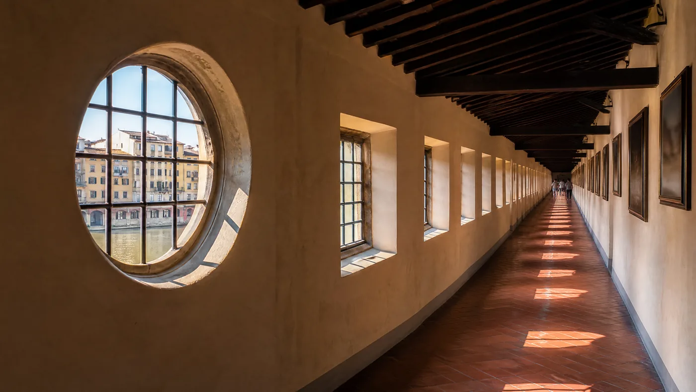 Un lungo corridoio stretto con finestre basse e una finestra rotonda sull'Arno